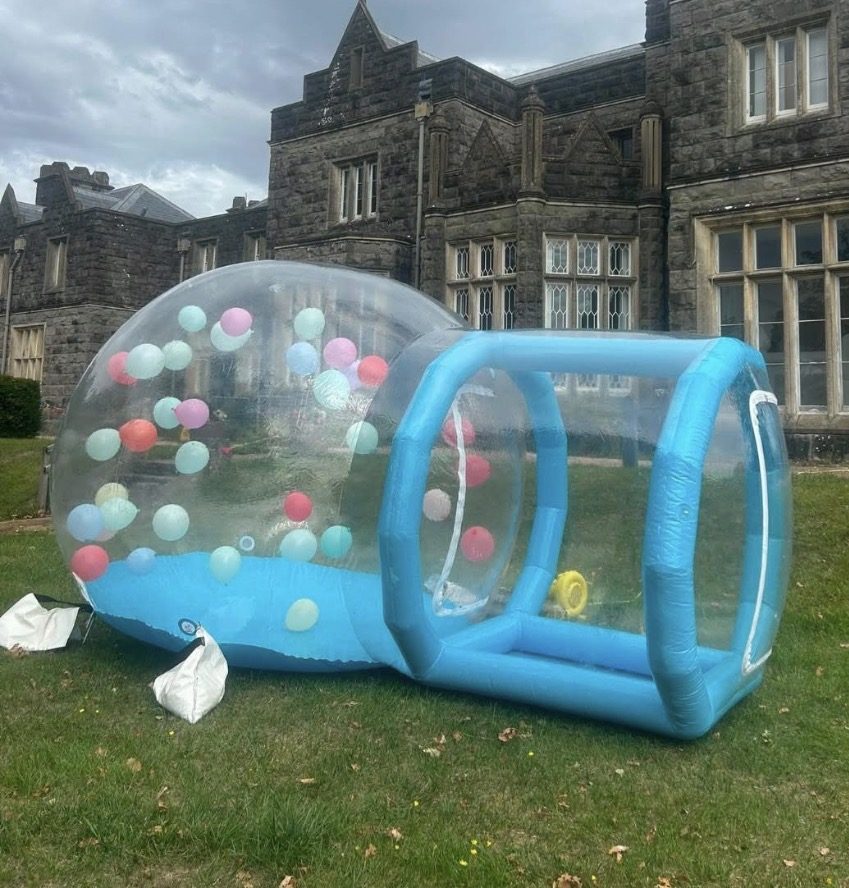 Blue and clear inflatable bubble house with pastel balloons in front of a stone building