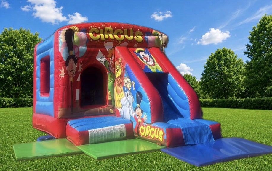Red and blue circus themed bouncy castle with a slide, clown and ringmaster artwork, on a lawn