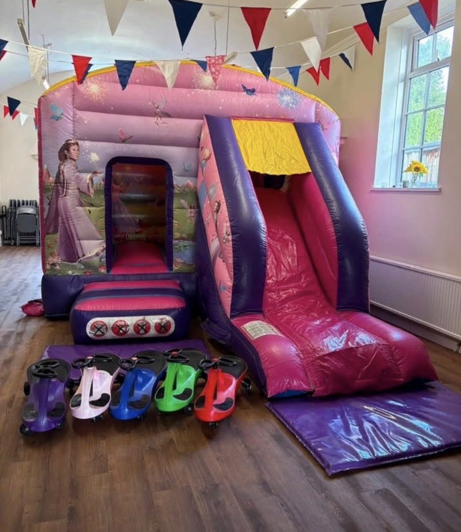 Pink and purple fairy princess bouncy castle with slide, set up in a hall with bunting and ride-on cars