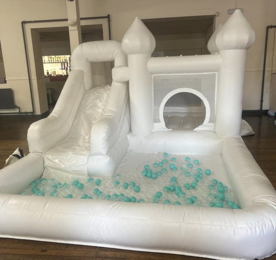 White bouncy castle with slide and ball pit filled with mint and clear balls, set up in a hall