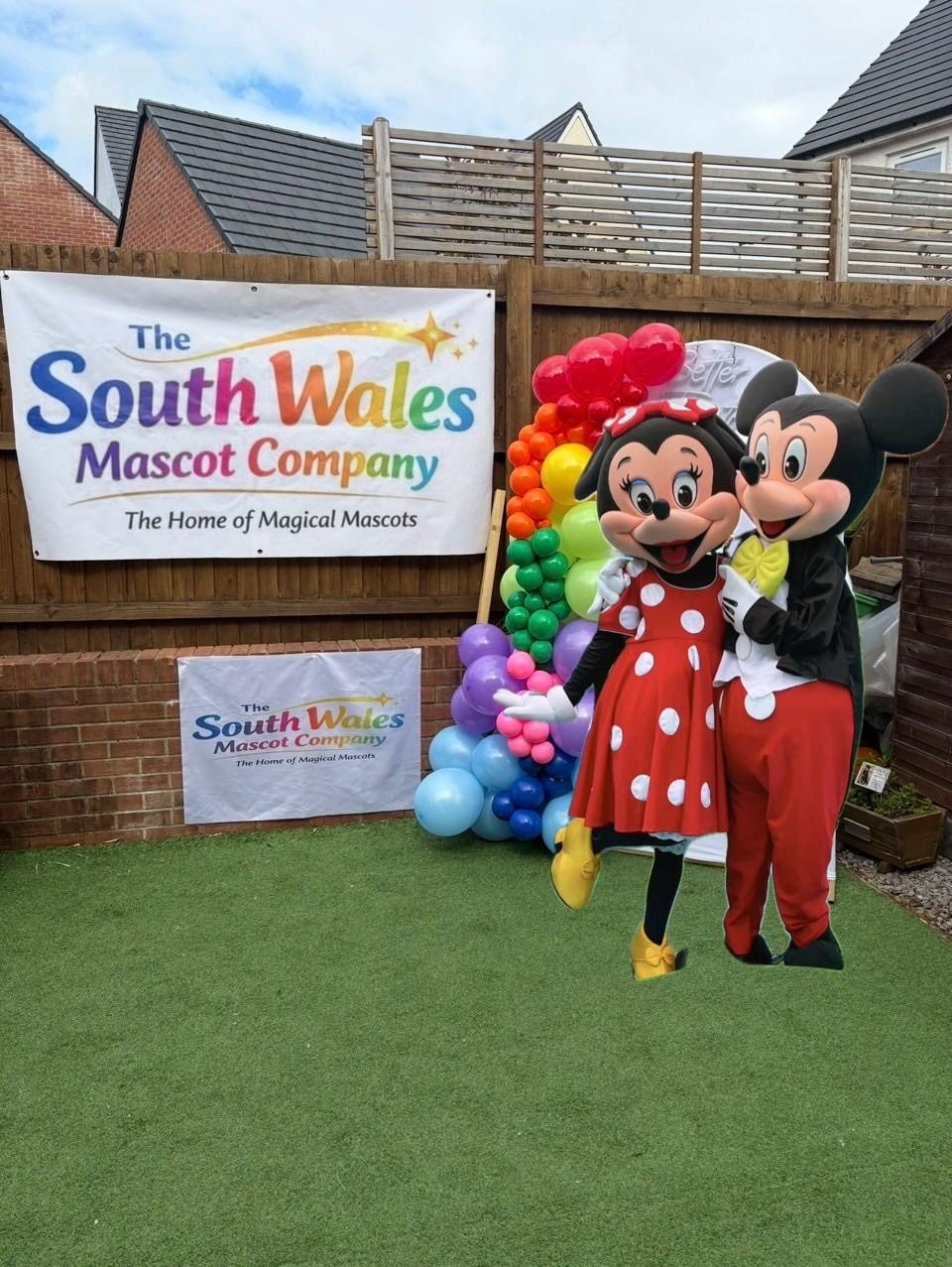 Mickey and Minnie Mouse lookalike mascots posing together by a rainbow balloon arch