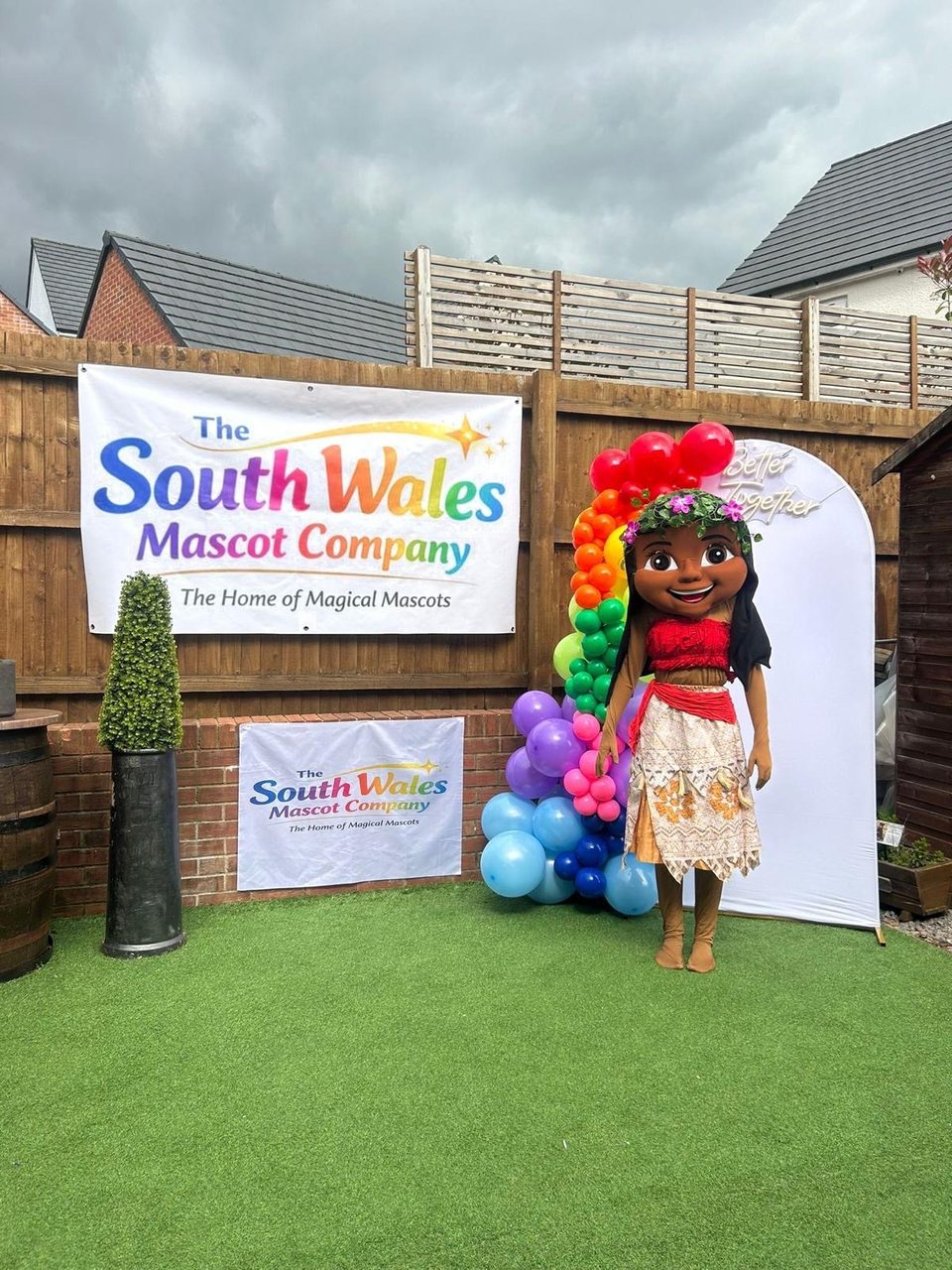 Moana lookalike mascot with a flower crown beside a rainbow balloon garland