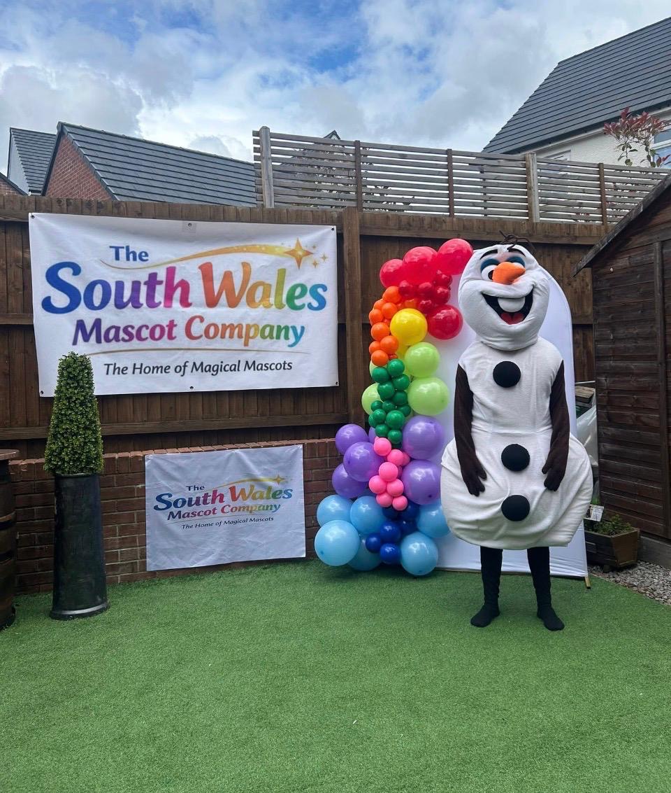 Snowman lookalike mascot beside a rainbow balloon garland and South Wales Mascot Company banners